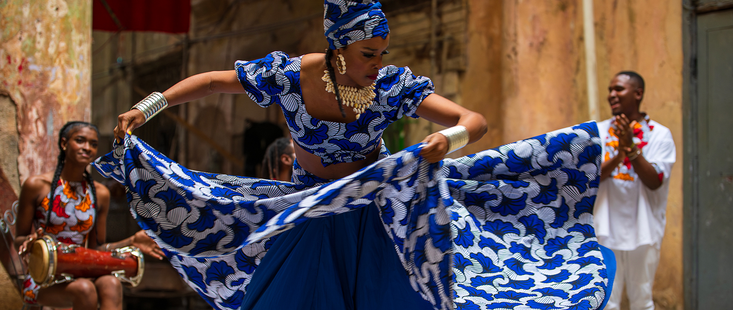 A person dances energetically in a vibrant blue and white traditional outfit on a street.