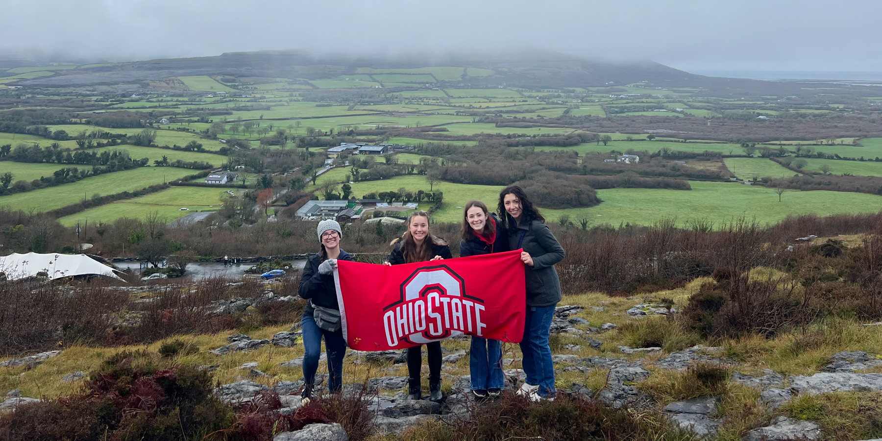 Four individuals holding an Ohio State banner in a scenic countryside with green fields and cloudy skies.