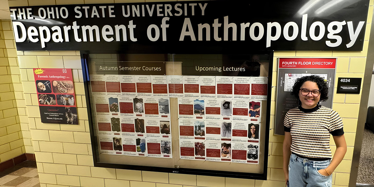 A person standing next to a bulletin board labeled "The Ohio State University Department of Anthropology," which displays notices about courses and lectures.