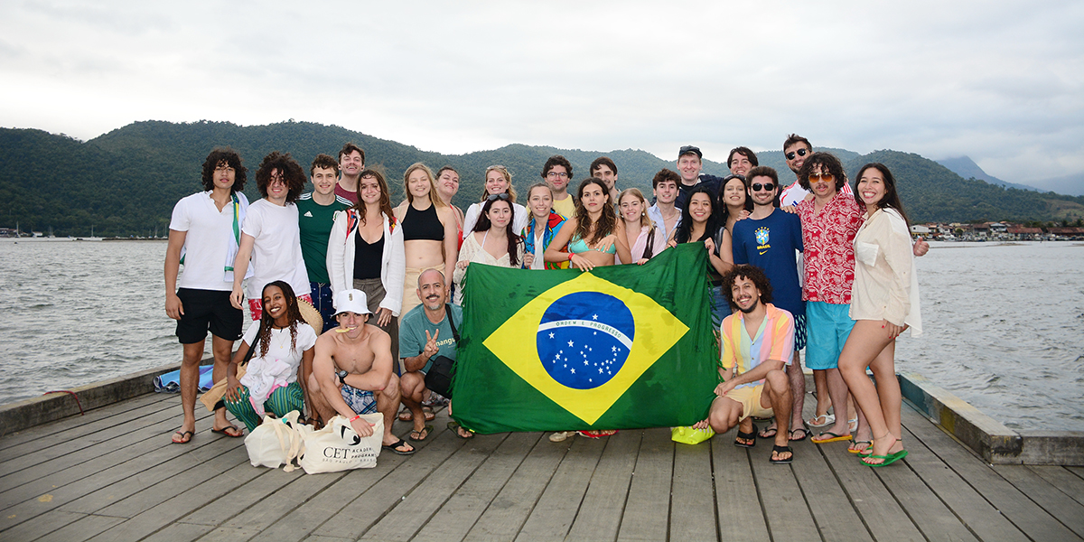 Group of people holding a large Brazilian flag, standing on a wooden pier with mountains and clouds in the background.