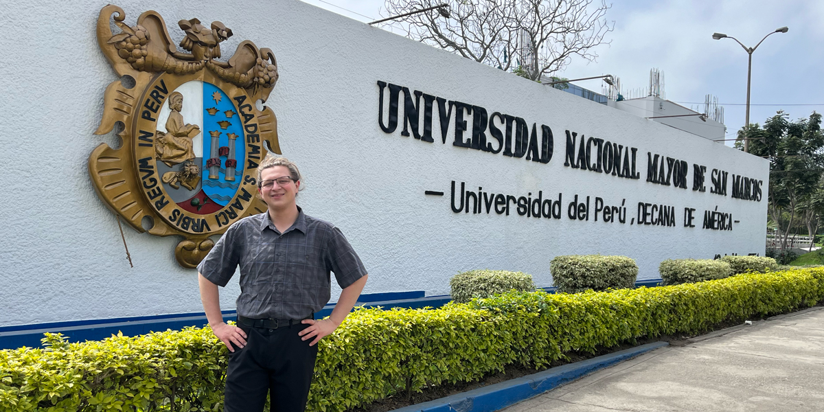 Person standing in front of the Universidad Nacional Mayor de San Marcos sign, with crest and landscaping visible.