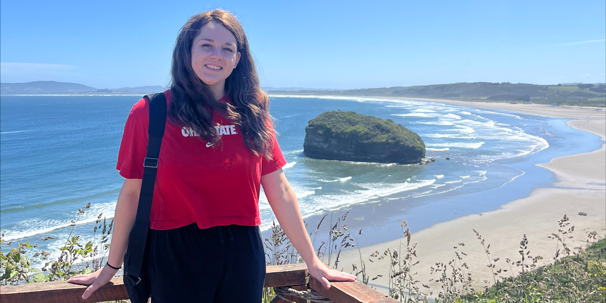 Person smiling at the camera, standing on a wooden overlook with a scenic view of a sandy beach and large rock formation in the ocean, under a clear blue sky.