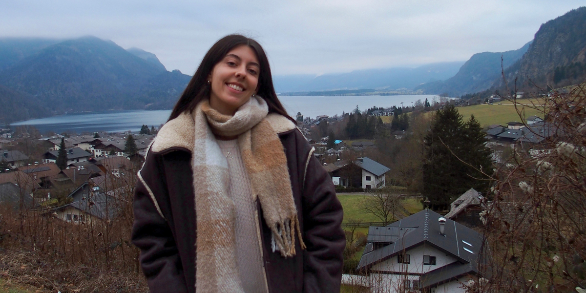 Person smiling, wearing a scarf and jacket, standing before a scenic view of Hallstatt village and lake in Austria.