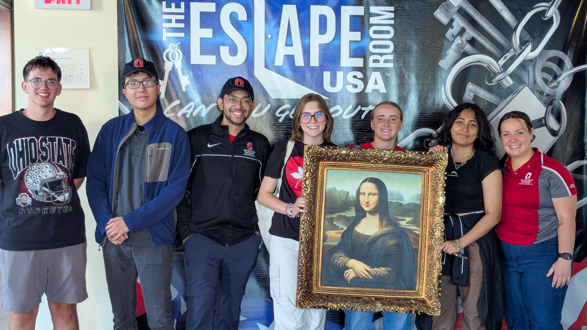 Seven people wearing Ohio State clothing at indoor setting, three holding a Mona Lisa painting.