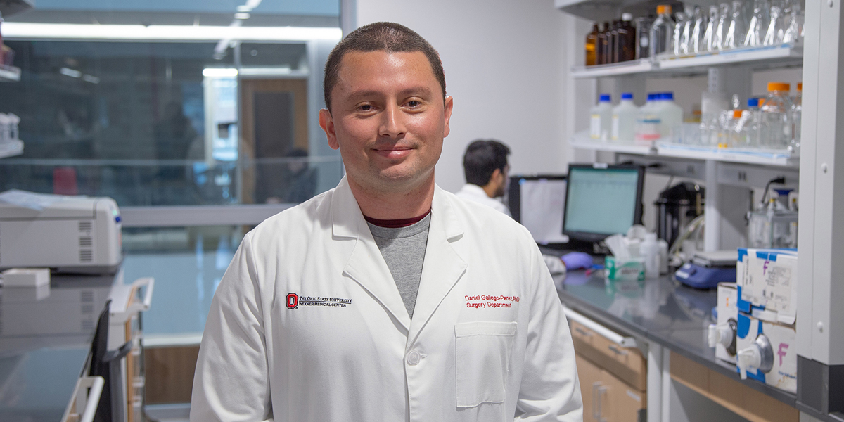 A person in a lab coat stands in a scientific laboratory setting. Shelves with bottles and equipment are in the background.