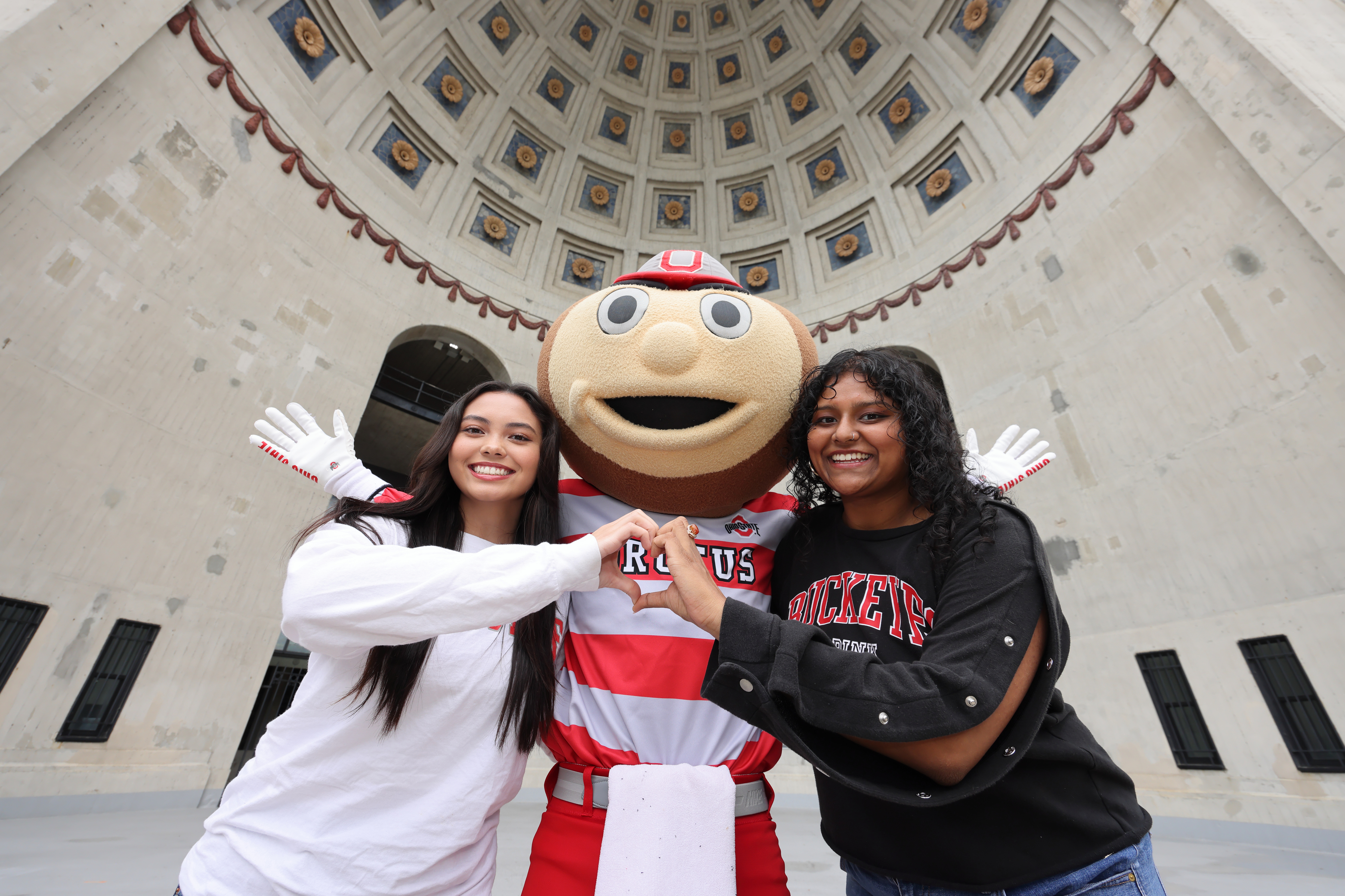 Two young people making a heart with their hands in front of the Brutus Mascot