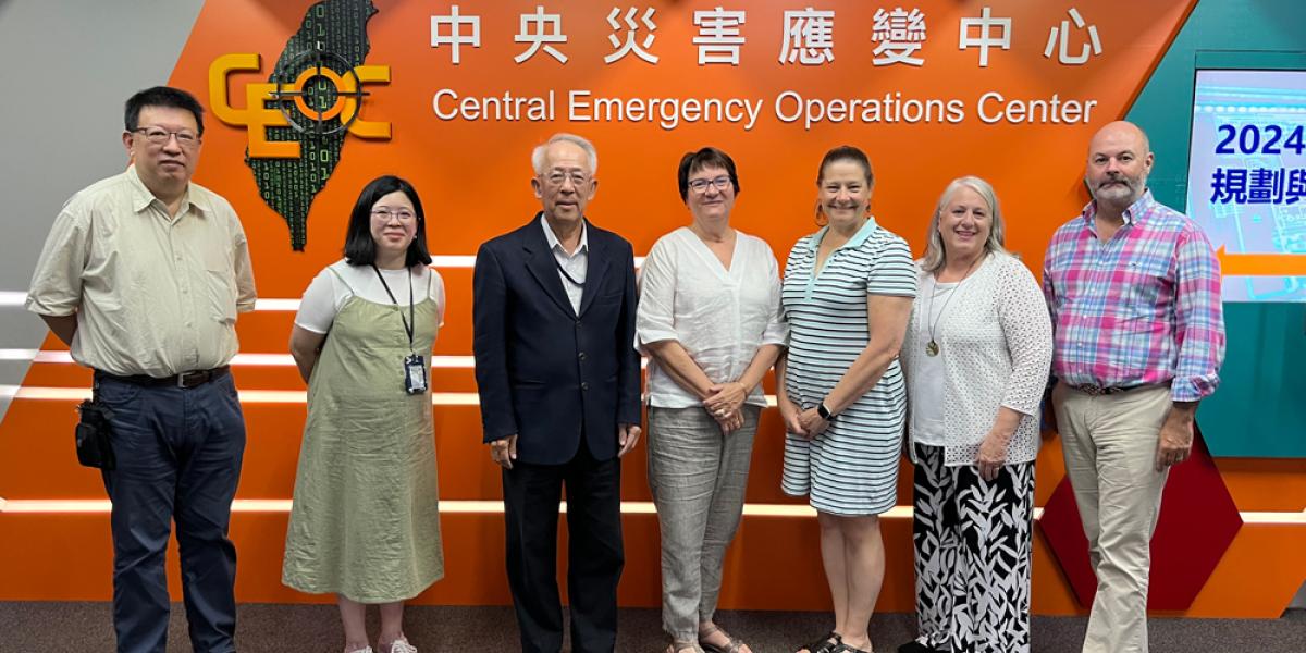 Group of seven adults standing in front of a sign that reads “Central Emergency Operations Center” with a digital map of Taiwan in the background.