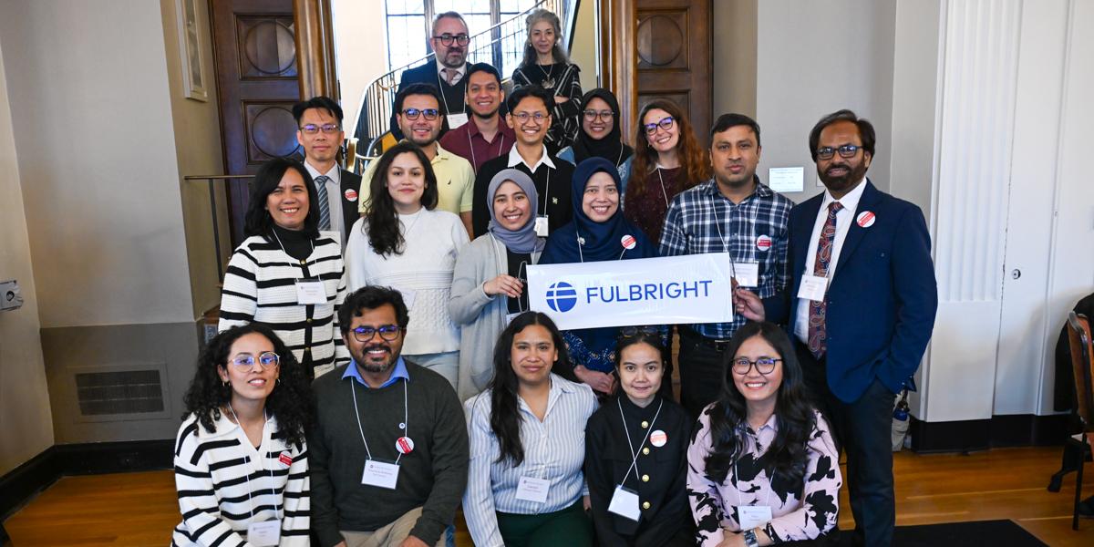 Large group of students and scholars gather indoors with a Fulbright sign.