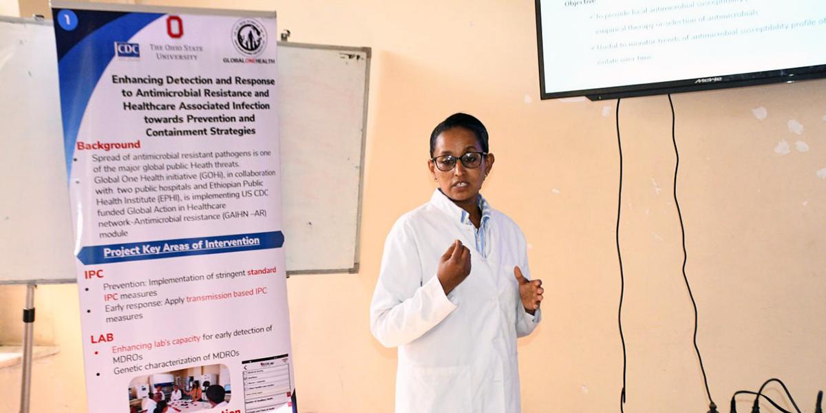 A woman presenting in front of a banner and monitor.