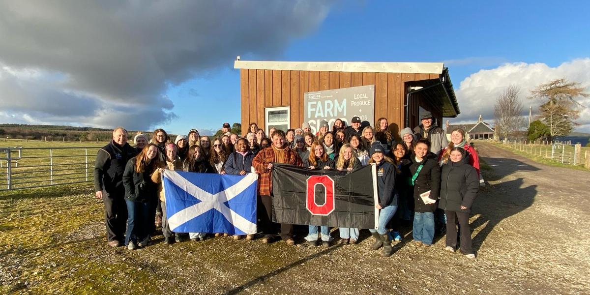 Large group of students holding Scotland flag and Ohio State flag pose in front of a barn.