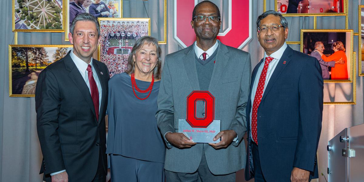 Three men and one woman in formal suits at award reception.