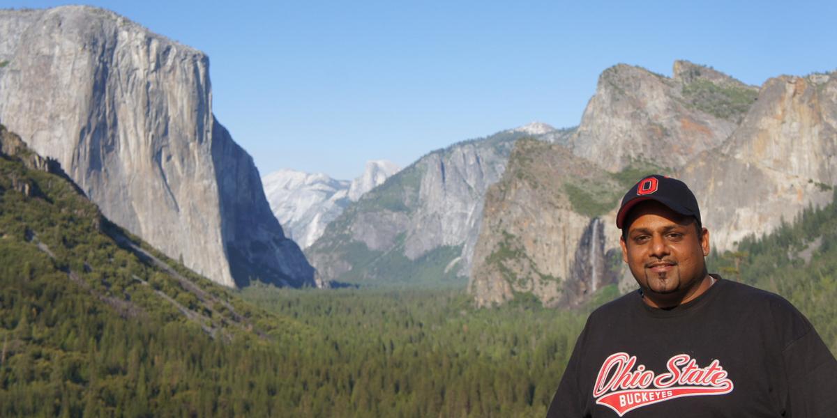 Person wearing an Ohio State Buckeyes shirt and cap, standing in front of Yosemite National Park's iconic El Capitan and Half Dome cliffs.