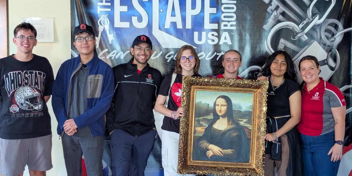 Seven people wearing Ohio State clothing at indoor setting, three holding a Mona Lisa painting.