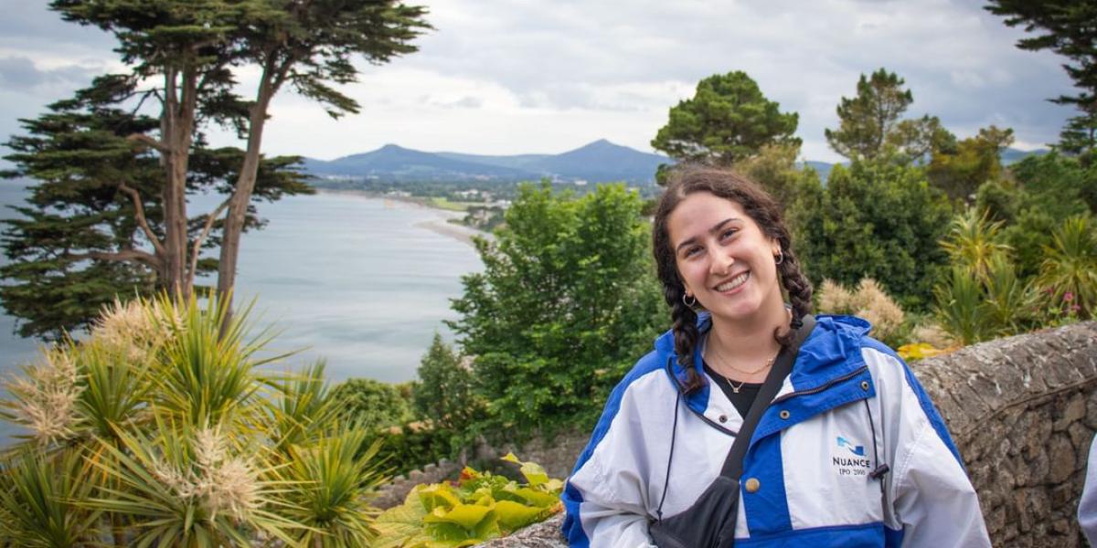 Young woman with brown hair in a windbreaker poses in front of an Irish landscape