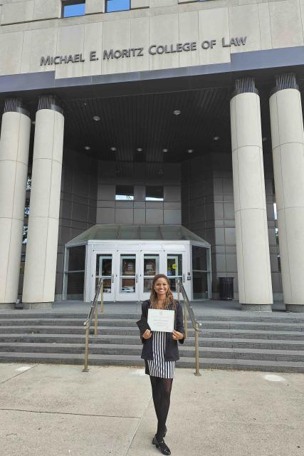 Suélen Pereira standing with her certificate in front of the Moritz College of Law.