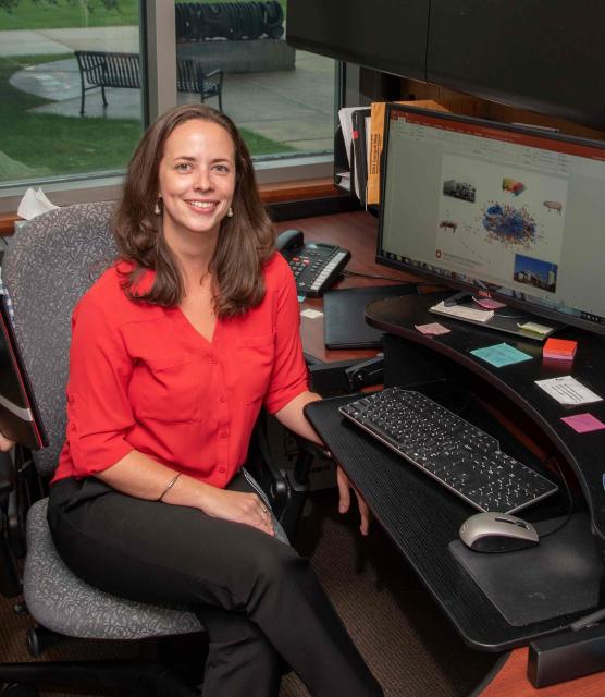 A person in a red shirt smiling at the camera, seated at a desk with a computer displaying a scientific graph, in an office environment with a view of a lawn through a window.
