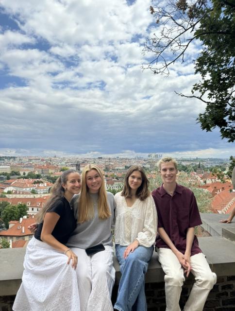 Three women and one man sit on a wall with city in background.