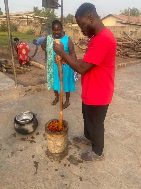 A person is using a large wooden pestle to pound food in a mortar, with another person standing nearby, observing the process. There is a cooking pot on a fire in the background, and someone else is seen engaging in activities behind them.