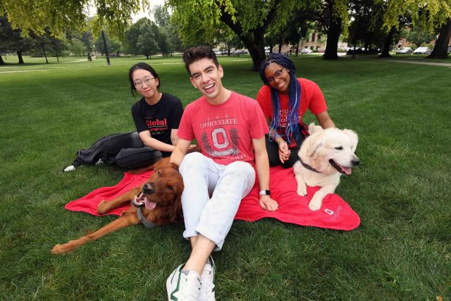 Three students sit on the Oval with dogs from Buckeye Paws.