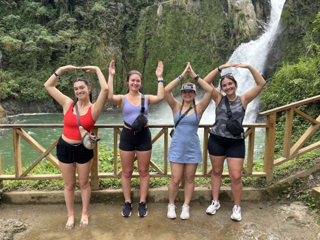Four people spelling out O-H-I-O with their arms raised in front of a waterfall.