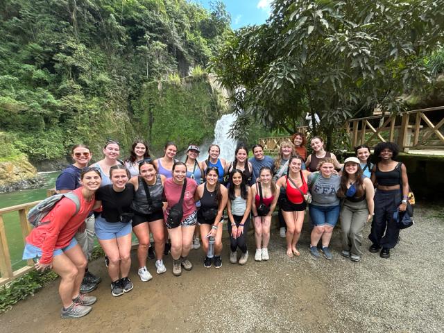 Group of students posing for a photo in front of a waterfall and a wooden bridge at a scenic outdoor location.