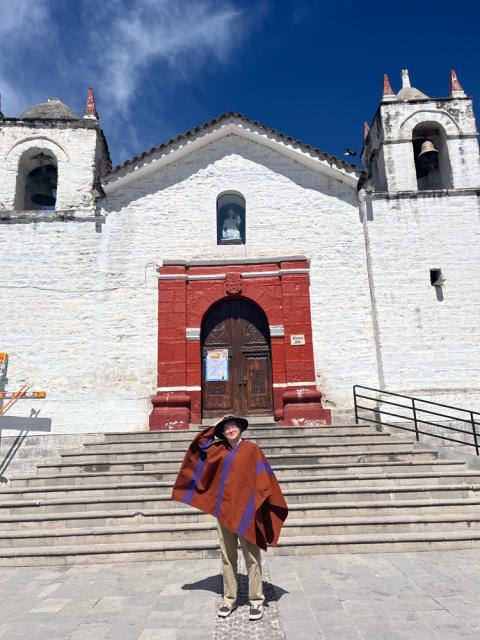 A person wearing a poncho stands smiling on the steps of San Pedro Claver Church in Cartagena, Colombia, under a clear blue sky.