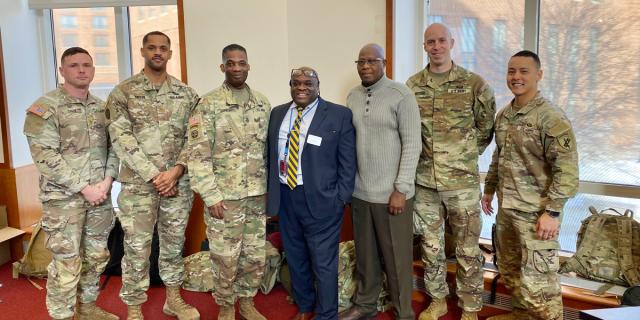Group of five military personnel in camouflage uniforms standing with two civilians in an office setting.