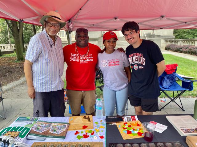 Four people standing under a red tent at an outdoor event, smiling at the camera wearing casual clothing.