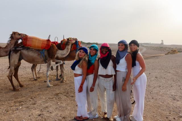Group of five people wearing traditional headscarves and white outfits, standing next to a camel in a desert setting.