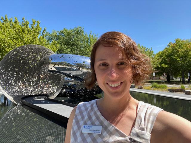 Person with a name tag smiling in front of the Canberra Times Fountain on a sunny day.