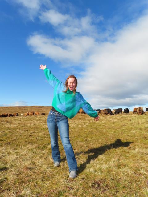 Woman with arms outstretched in a field with cows behind her