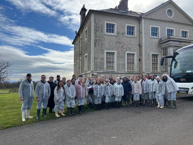 Large group of students wearing hazard suits pose by farm house.