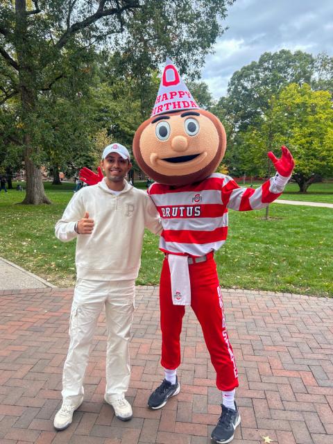 An individual in a white hoodie posing next to Brutus Buckeye, who is wearing a red and white outfit and a birthday hat, on a tree-lined pathway.