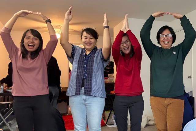 Four people standing side by side, smiling and forming O-H-I-O with their arms raised overhead. Each person wears colorful attire.