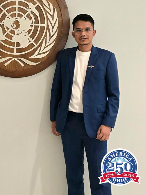 Man wearing a blazer and glasses stands by UN sign.
