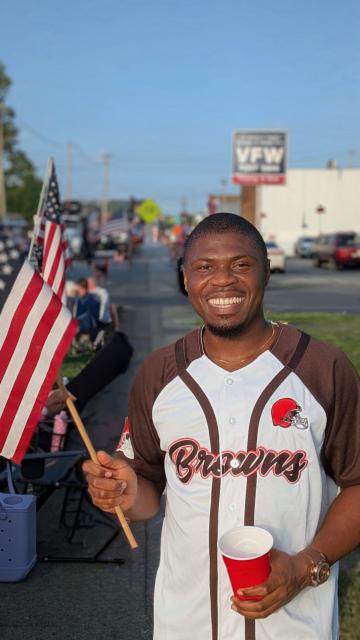 A man smiling and holding an American flag is wearing a sports jersey with "Browns" and the Cleveland Browns logo outdoors on a sidewalk during an event.