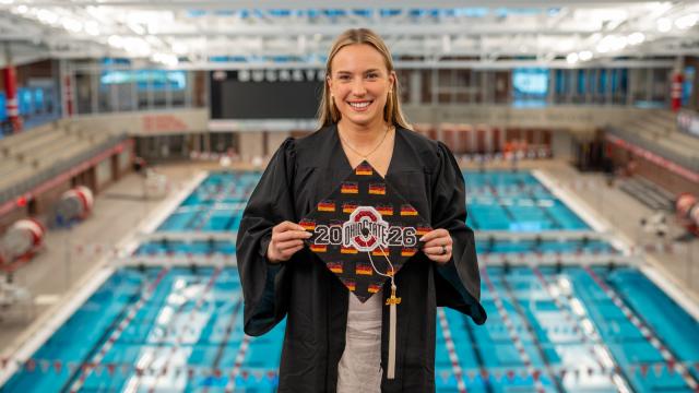 Female wearing graduation gown holding grad cap with Ohio State logo with pool in background.