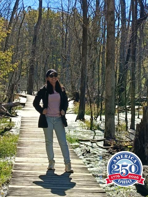 Woman wearing a jacket, hat and sunglasses stands on a wooden trail.
