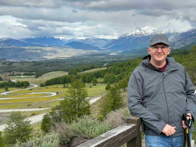 A person stands on a wooden deck overlooking a scenic landscape in Patagonia. The view includes winding roads, lush green fields, trees, and distant snow-capped mountains under a cloudy sky. The person wears a gray jacket and cap, holding a walking stick, and smiles at the camera.