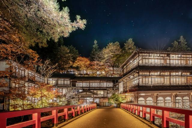 Night view of a traditional Japanese inn surrounded by trees, with a red bridge leading to the entrance.