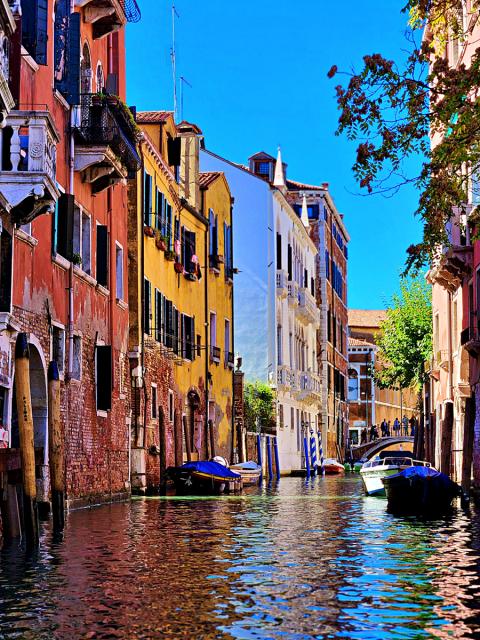 A vibrant canal in Venice, Italy, flanked by colorful buildings with arched windows. Several small boats are tied along the water, and blue-and-white striped mooring poles are visible. 