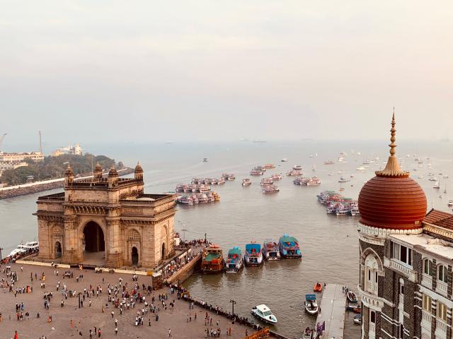 View of the Gateway of India and the Taj Mahal Palace Hotel by the waterfront in Mumbai. Boats are visible in the harbor