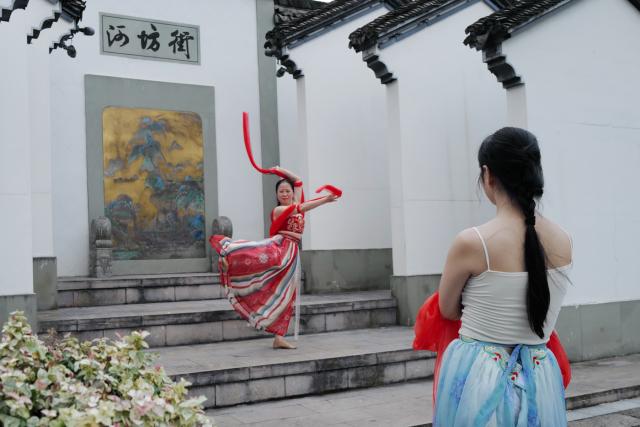 A person in a colorful dress performs a traditional dance with red ribbons in a courtyard, as another person watches. Chinese architectural elements are visible/