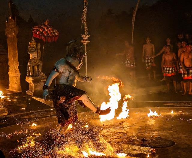 A performer in traditional attire dances through fire during a ceremonial event at night. The scene is illuminated by flames, with statues and a group of onlookers in the background