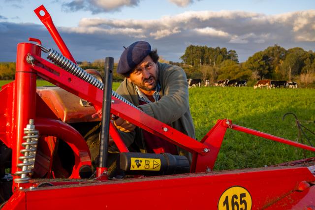 A person is working on red agricultural machinery in a green field. A herd of cows grazes in the background, with trees and a cloudy sky visible.