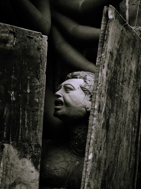 A dark, moody sculpture of a person with an expressive face, partially obscured by rugged wooden planks. The background features twisting, pipe-like structures.