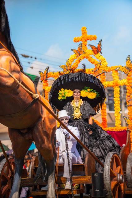 A vibrant scene from a Día de los Muertos parade featuring a person in an elaborate black costume and large hat adorned with yellow marigolds, riding a wooden cart