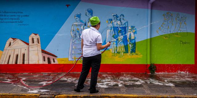 A person cleans a colorful mural depicting various characters and symbols, including historical figures and cultural motifs, on an urban wall.