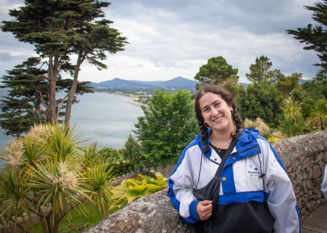 Young woman with brown hair in a windbreaker poses in front of an Irish landscape