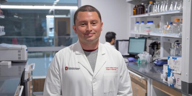 A person in a lab coat stands in a scientific laboratory setting. Shelves with bottles and equipment are in the background.
